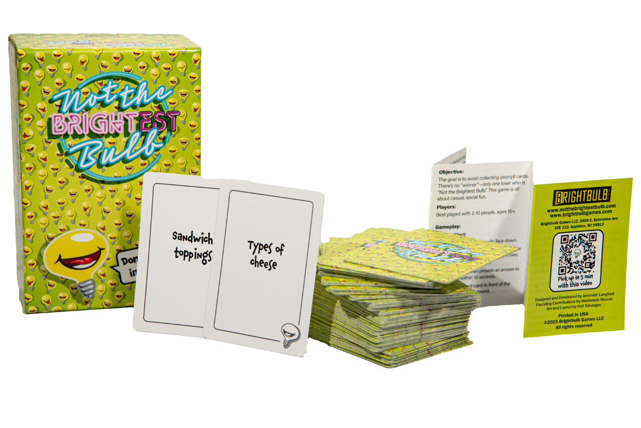 Photo of the full “Not The Brightest Bulb” game set, including the box, prompt cards like “Sandwich toppings” and “Types of cheese,” and the rulebook with QR code.