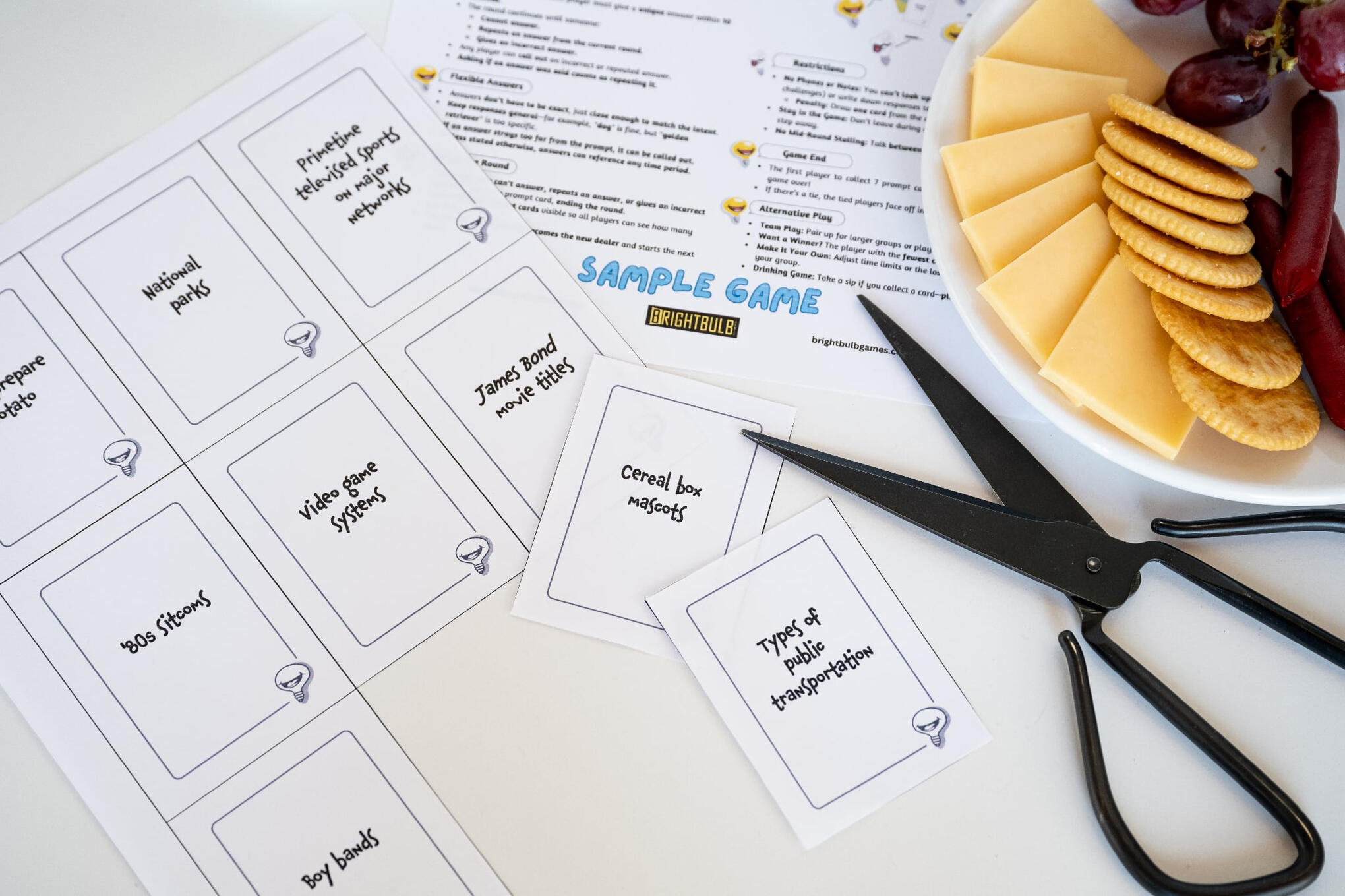 Print-and-Play Sample with Snacks and Scissors Top-down photo of the print-and-play sample game for Not The Brightest Bulb, with uncut black-and-white cards labeled with prompts like "Types of transportation" and "Cereal box mascots," plus scissors and a snack plate with cheese and crackers